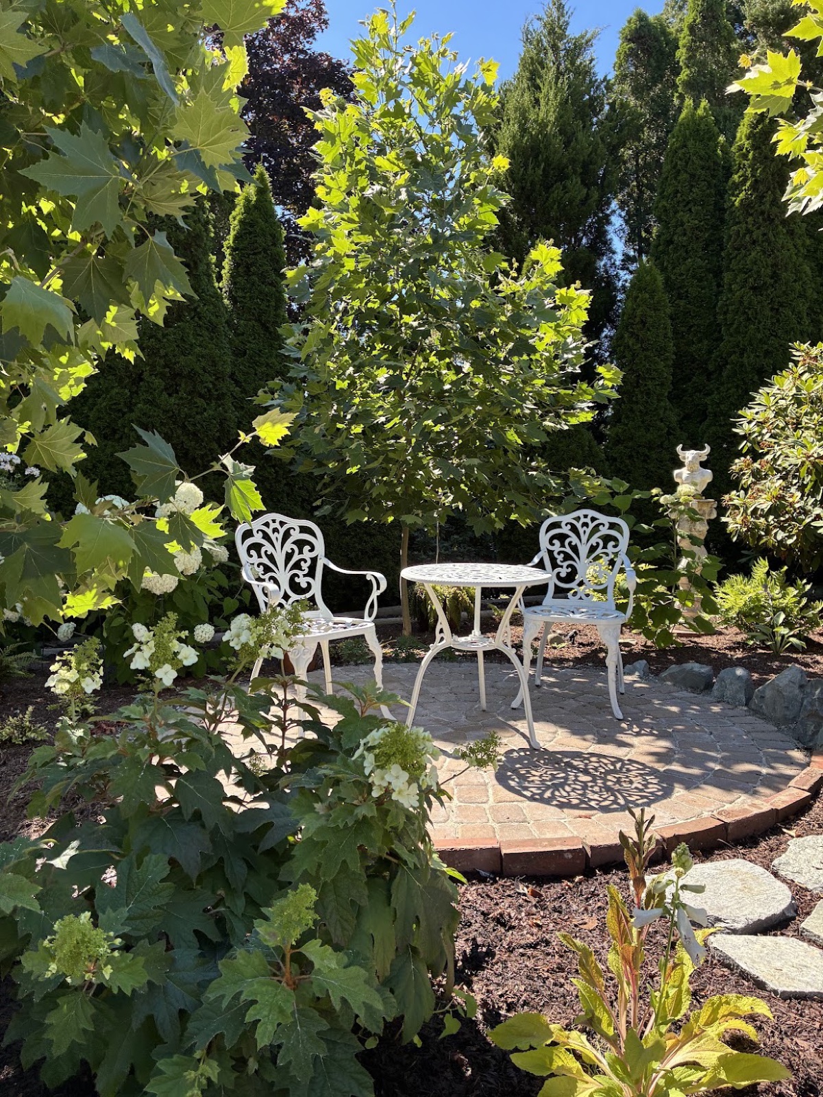 A white cast-iron bistro table and two chairs on a circular brick patio, ringed by London plane trees, arborvitae, and white hydrangeas in summer sun.